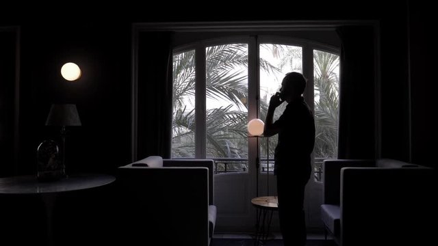 Silhouette of man in a dark room next to a day window, talking on the smart phone, very angry
