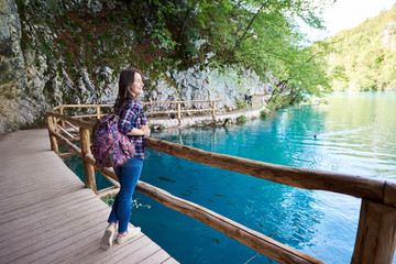 Slim pretty happy tourist woman with backpack standing on wooden pathway under green tree enjoying beautiful view of clean blue lake lit by summer sun. Plitvice National Park, Croatia, Europe.