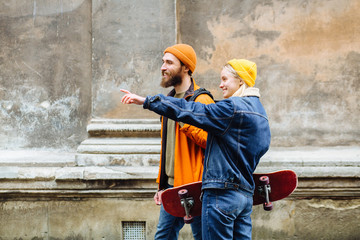Happy young hipster couple traveler hold hands and walking together with skateboards over old wall on street in windy cold rainy weather outdoor.