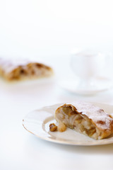 Apple strudel served on a white porcelain plate. Coffee cups in bokeh, white table, high resolution