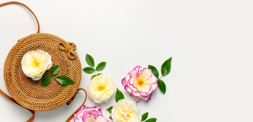Fashionable concept of women's handbags. Handmade natural organic rattan bag and delicate rose flowers with green leaves on light background. Flat lay, Copy space, top view. Ecobags from Bali.