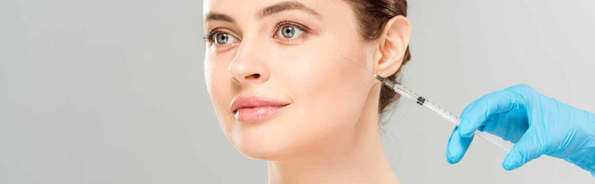 panoramic shot of cosmetologist in blue latex glove holding syringe near face of attractive woman isolated on grey
