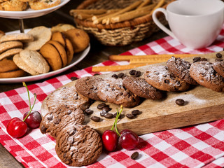 Oatmeal chocolate cookies with coffee grains and cherry, powdered sugar on kitchen cutting board with checkered fabric on wooden table in village style for picnic. Breakfast for loved one.