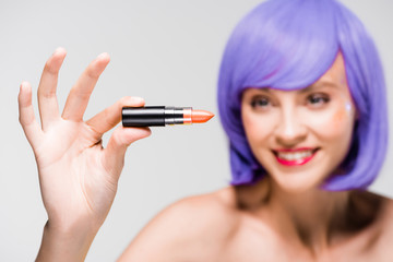 selective focus of beautiful young woman in purple wig holding lipstick isolated on grey