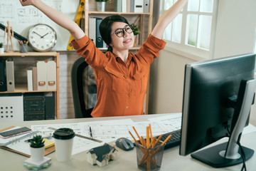 Happy young emotional entrepreneur feeling excited and happy of working process making job great indoors. successful smiling woman architect in spectacles sitting at desktop stretching arms joyful