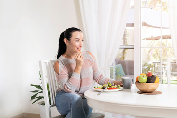 Obraz na płótnie Canvas Beautiful brunette woman sitting at dining table eating a healthy salad and drinking coffee or tea with a bowl of fruits on table with garden in the background