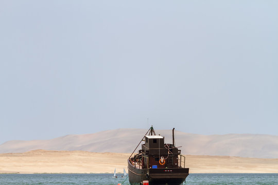 Old wooden boat sails the coast of Paracas (Peru) with dunes in the background