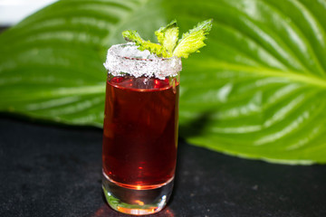A glass is sprinkled with sugar, there is a strawberry tincture inside, a sprig of mint on top against a dark background, and the green leaves of the Hosts are in the background.
