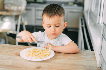 little boy eats pasta in the form of a spiral in the afternoon in the kitchen on their own