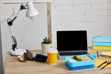 blue plastic lunch box with healthy food on wooden table with laptop and succulent on white background, illustrative editorial
