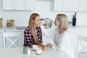 Two attractive lesbian female in casual clothes sitting together at the table in the kitchen, looking at each other