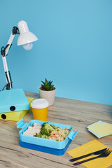 lunch box with broccoli, rice, chicken and coffee in plastic cup at workplace on wooden table on blue background