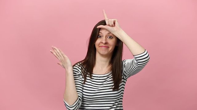Smiling pretty young woman in striped shirt, fooling, showing loser gesture, pointing index finger on camera isolated over pastel pink background in studio. People sincere emotions lifestyle concept.