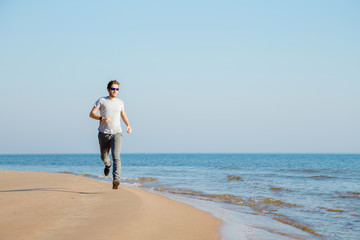 Fototapeta na wymiar Young, adult alone man in t-shirt, sport trousers and sunglasses running on sand beach. Empty place for motivational, inspirational text, quote or sayings on light blue sky background. Front view. 