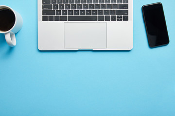 Top view of laptop keyboard, computer mouse, smartphone and cup of coffee on blue background, illustrative editorial