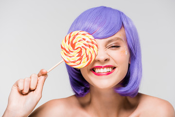 cheerful girl in purple wig holding sweet lollipop, isolated on grey