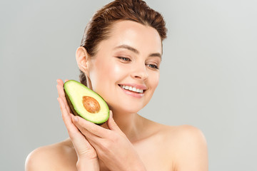 happy and naked woman holding half of ripe avocado isolated on grey