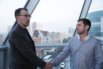 Two businessmen shaking hands in front of a panoramic office window through which a street with office buildings, city buildings and cars is visible. With copy space fo text and image.