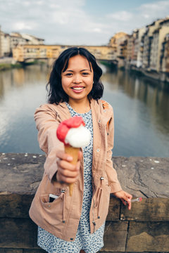 Young Filipino woman shows her ice cream with back Ponte Vecchio. Summer in the city, teenager with the cone on the Arno River. Historic symbol of the city of Florence, the old bridge