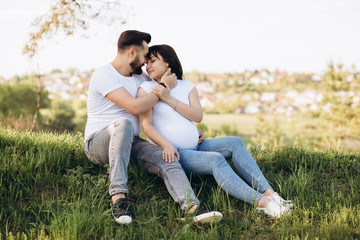 Happy young couple expecting baby, pregnant woman with husband touching belly, sitting on green grass