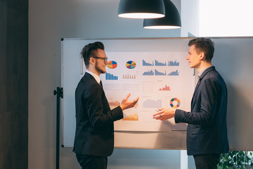 Corporate meeting. Two young men standing at the whiteboard with graphs, discussing startup business strategy.