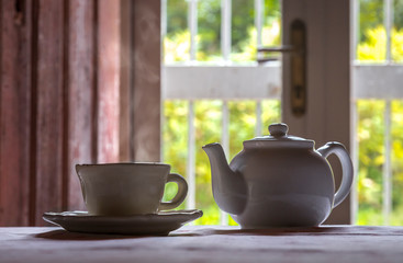 Una taza de té humeante junto con una tetera blanca con un fondo de puerta y luz día