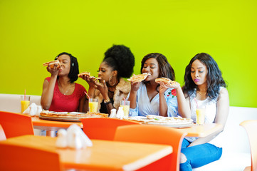 Four young african girls in bright colored restaurant eating pizza slices in hands.