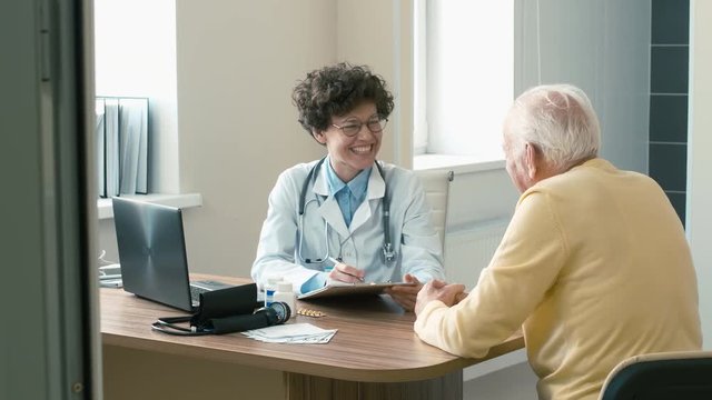 Female doctor's room where she consulting old grey-haired man, smiling, asking about health and doing some notes
