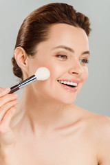 Fototapeta premium cheerful woman applying blush on cheek with cosmetic brush and smiling isolated on grey