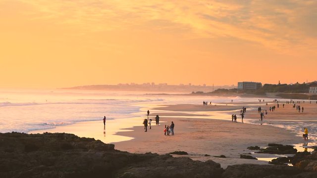 Slow motion of people at Carcavelos beach, Slowly waves form and break, beautiful Atlantic ocean, deep yellow sunset on water reflections, Portugal