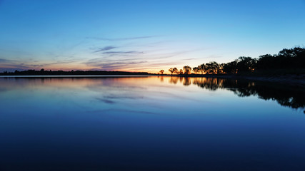 Sunset overlooking the lake, trees, clouds. The sky and bushes are reflected in the water. Landscape at dusk. Nightfall. Water like a mirror.