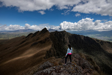 Young girl on top of a mountain looking at the horizon, in the background three mountain peaks with a blue sky and some banks of clouds