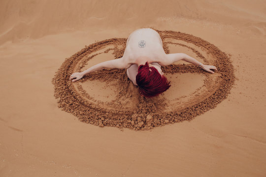 Naked redhead girl drawing sign on sand with her hands