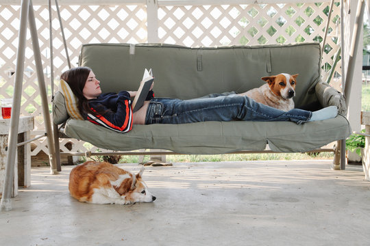 young woman reads book while relaxing in patio swing with dogs