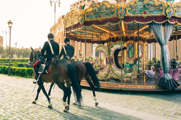 Couple of police on horseback passing by a carousel in the city of Rome. Warm, soft and orange...