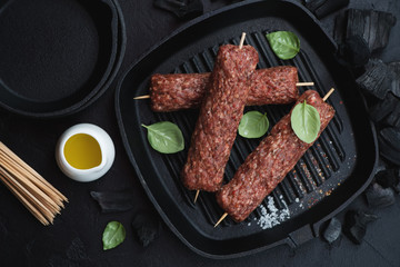 Raw marbled beef skewers on a cast-iron grill ready to be cooked, flatlay over black stone background with charcoal