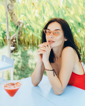 the girl in pink glasses and a bathing suit sits at a table and drinks a water-melon juice