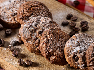 Oatmeal chocolate cookies with coffee grains, powdered sugar on kitchen cutting board with checkered fabric on wooden table in village style for picnic. Dark environment. Served hot.