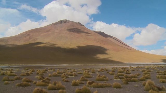 Wide low-angle panning shot of Andean desert landscape with brown bare mountains, dry yellowish grass, and a blue sky with scattered white clouds, Isluga River, Bolivia