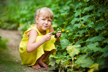 The smiles  girl in a yellow dress sits in a kitchen garden and holds a cucumber in hand
