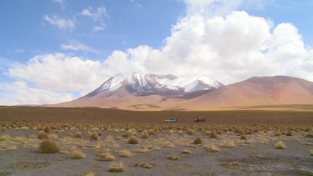 Wide, low-angle zooming shot of altiplano desert landscape with a snow peaked andean mountain and arid vegetation. Two 4x4 off-road vehicles are parked with tourists enjoying the altiplano scenery