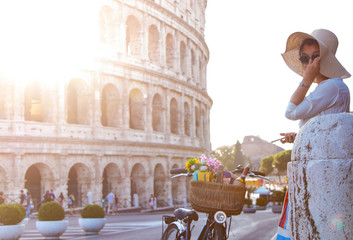 Beautiful young woman with bike in fashion dress standing in front of colosseum in Rome at sunset.