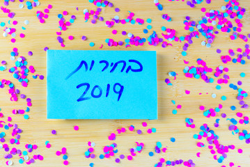 Hebrew text Elections 2019 on voting paper over wooden board with confetti background. Israel new Elections 17 September 2019.