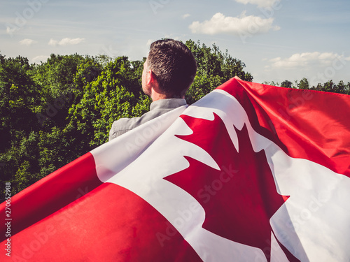 Handsome man waving a Canadian Flag against a background of trees and blue sky. View from the back, close-up. National holiday concept