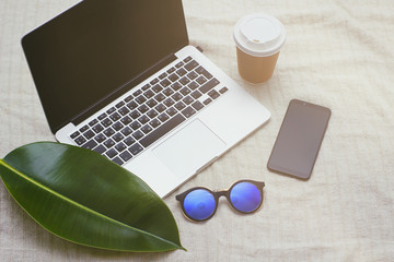 Fototapeta na wymiar Open laptop on home cloth background. Paper disposable cup of coffee, black mobile phone, blue sunglasses, big exotic leaf. Online work concept.