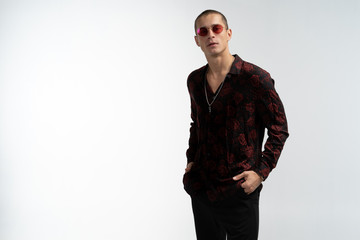 Fototapeta premium Full length portrait of amazing attractive man in stylish trendy clothes, wearing red sunglasses, holding hands in pockets, looking at camera.