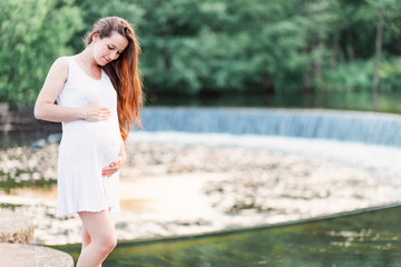 Beautiful pregnant woman stands near the river with a small waterfall
