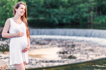 Beautiful pregnant woman stands near the river with a small waterfall