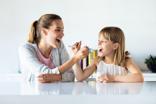 Beautiful mother and daughter feeding yogurt to each other at home.