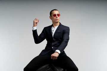 Attractive young man in black suit and red sunglasses sitting on the big tire in the pose of a rider, isolated over white background